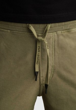Close-up van olijfgroene joggingbroek met zwart afgewerkte trekkoorden, gestrikt in een strik, met de tekst "PME Legend" op de koorden.
