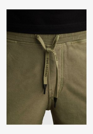 Close-up van olijfgroene joggingbroek met zwart afgewerkte trekkoorden, gestrikt in een strik, met de tekst "PME Legend" op de koorden.