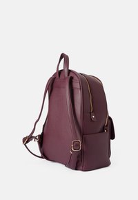 Mauve plecak ze sztucznej skóry o strukturalnym kształcie, złotymi elementami, podwójnymi zamkami błyskawicznymi oraz boczną kieszenią. Regulowane paski z uchwytem u góry.