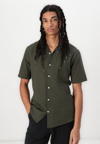 Camisa de manga corta, de color verde oscuro, hecha de un tejido ligero con botones en la parte delantera y un cuello puntiagudo, que presenta un dobladillo curvado.