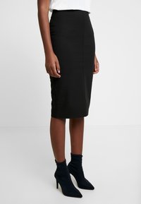 Jupe crayon noire taille haute, coupe ajustée et texture lisse. Associée à des bottines pointues bleu marine à enfiler avec un haut souple.