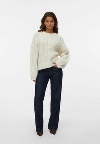 Weißer Strickpullover mit einem strukturierten Kabelmuster, kombiniert mit dunkelblauen geraden Jeans und spitzen braunen Schuhen.
