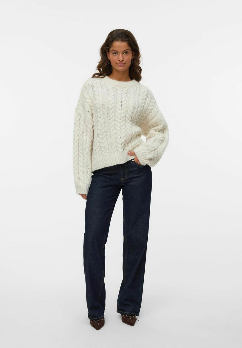 Weißer Strickpullover mit einem strukturierten Kabelmuster, kombiniert mit dunkelblauen geraden Jeans und spitzen braunen Schuhen.