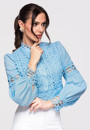 Femme aux cheveux foncés en queue de cheval portant de grandes boucles d'oreilles florales et un chemisier en dentelle bleu clair avec des détails découpés sur les manches et à l'avant.