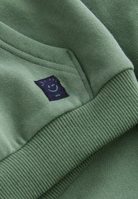 Sweat-shirt vert avec des poignets et un ourlet côtelé, orné d'une étiquette en tissu bleu foncé affichant un emblème de visage souriant près de la poche.
