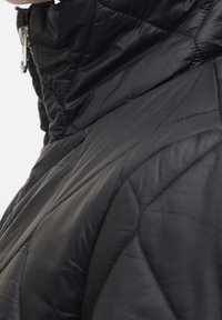 Veste noire matelassée avec un col montant, présentant des coutures diagonales et une fermeture éclair. Conçue en matériau léger et lisse.