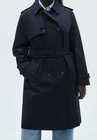 Osoba oblečená v černém trenčkotě s páskem a dvojitými knoflíky, pod ním má světle modrou košili a džíny, ruce v kapsách kabátu.