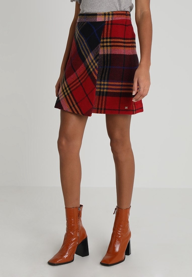 Femme portant une mini-jupe à carreaux rouge et noir et des bottines marron brillantes à talons épais, debout devant un fond uni.