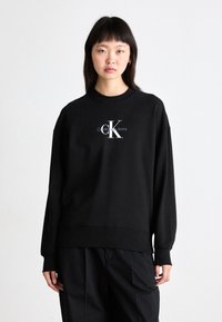 Črna bombažna majica s krožnim vratom in rebrastimi manšetami, ki na sprednji strani prikazuje belo in modro "CK Calvin Klein Jeans" logo.