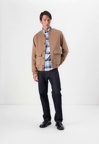 Cardigan marron texturé avec boutons et poches avant, superposé à une chemise à carreaux bleue. Porté avec un jean foncé et des chaussures noires.