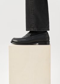 Svarta lädermockasiner med en slät yta, med sömmar som detaljer, och en låg klack, bärs med mörka denimbyxor på en ljus pelare.