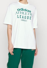 Valkoinen puuvillapaita, jossa on vihreä painatus "adidas ATHLETIC LEAGUE" ja logo. Ominaisuuksia ovat lyhyet hihat ja rento istuvuus.