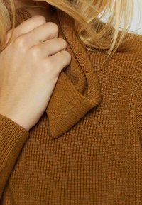 Pull en maille brune et texturée avec un col plié. Une main tient le col, mettant en valeur l'apparence douce et chaleureuse du tissu.