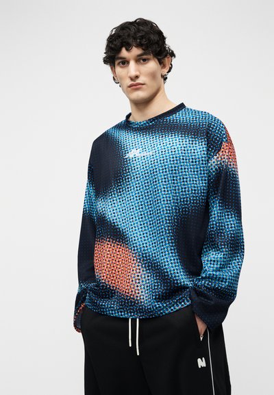 Jeune homme aux cheveux courts et bouclés portant une chemise à manches longues bleue et orange à motifs, et un pantalon noir avec des cordons blancs.