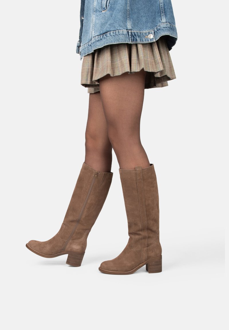 Mujer vestida con botas de ante marrones hasta la rodilla, medias transparentes marrones, una falda de cuadros y una chaqueta de mezclilla azul, de pie sobre un fondo blanco.