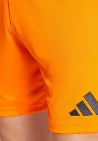 Oranssit urheilushortsit, jotka on valmistettu kevyestä, sileästä kankaasta; vasemmassa alakulmassa on musta Adidas-logo. Helma on suora ja siisti.
