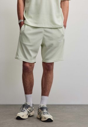 Mann trägt hellcremefarbene, strukturierte Shorts, weiße Socken und beige Laufschuhe, steht mit den Händen in den Taschen auf grauem Boden.