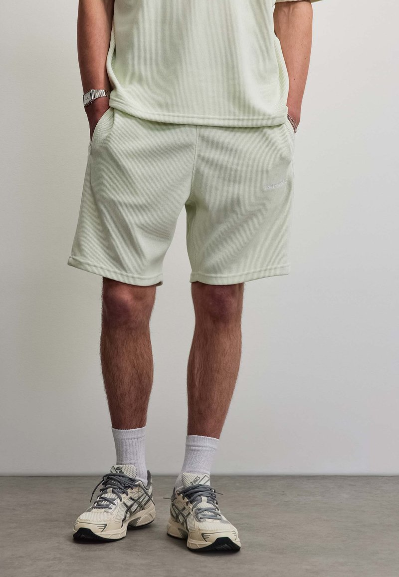 Homme portant un short texturé crème clair, des chaussettes blanches et des chaussures de course beiges, debout les mains dans les poches sur un sol gris.