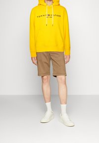 Sudadera amarilla con capucha y bolsillo frontal, con el logo "TOMMY HILFIGER". Combinada con pantalones cortos color caqui y zapatillas bajas blancas.