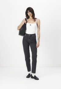 Balta bezpiedurkņu krekls ar melnu logo, augsta vidukļa tumšas džinsas, melna soma un melni loafers ar baltām zeķēm. Studijas vidē, vienkāršs balts fons.