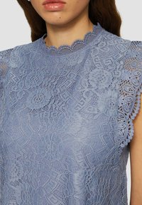 Jasnoniebieska bluzka koronkowa z falbaniastym dekoltem, kwiatowymi wzorami oraz krótkimi rękawami z misternymi detalami i wycięciami.