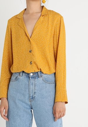 Nainen pukeutunut sinappikeltaiseen pitkähihainen puseroon, jossa on valkoisia pilkkuja, ja pusero on työnnetty vaaleansinisten, korkeavyötäröisten farkkujen sisään.