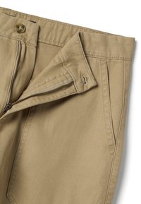 Pantalones beige con cremallera parcialmente abierta, botón y bolsillo frontal visibles sobre un fondo blanco liso.