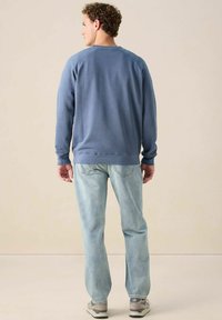 Un homme se tenant de dos, portant une chemise à manches longues bleue, un jean bleu clair et des baskets grises, sur un fond beige uni.