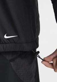 Svart Nike-jacka med strukturerad yta, elastisk nederkant och dragsko-detalj. Logotypen är tryckt i vitt på framsidan.