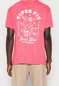 Person som bär en bleknad röd T-shirt med en vit orm som är hopkrupen runt ett skalle och texten "Viper Pit Sunset Blvd Los Angeles" på baksidan.