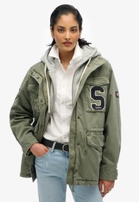 Oliwkowa kurtka z kapturem, zapinana na zamek, z przednimi kieszeniami i naszywką z literą "S". Noszona na białej koszuli i jasnoniebieskich dżinsach.