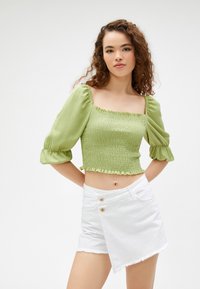 Grünes, gerafftes Crop-Top mit Puffärmeln und gewelltem Saum, kombiniert mit weißen High-Waist-Shorts im Wickeldesign und Knopfdetails.