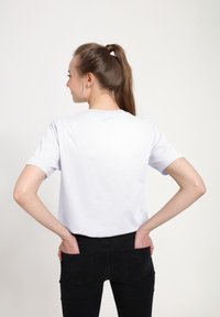 Bílý krátký rukáv tričko s kulatým výstřihem, zobrazené zezadu, v kombinaci s černými těsnými džíny, ruce v kapsách. Hladká textura, volný střih.