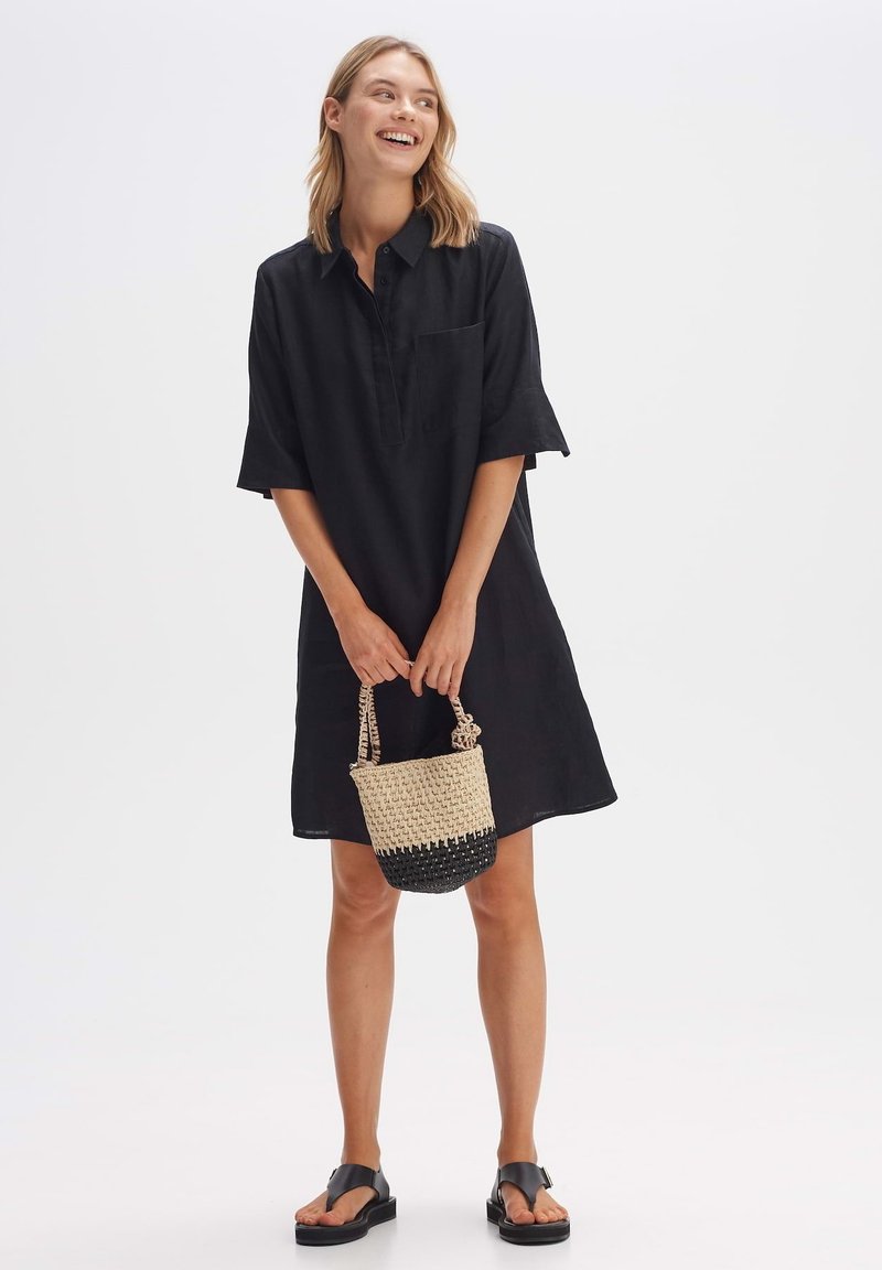 Zwart linnenhemdjurk met een voorzak en halve mouwen, gecombineerd met een geweven tas in zwarte en natuurlijke tinten, en platte zwarte sandalen.