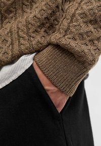 Brauner Strickpullover mit strukturiertem Muster, gerippte Bündchen, über einem hell gestreiften Hemd getragen, kombiniert mit schwarzen Hosen, Hand in der Tasche.