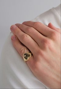 Inel auriu în formă de inimă cu un centru închis la culoare, accentuat de o mică piatră prețioasă, purtat pe o mână pe un material texturat, alb.