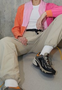 Veste bomber bicolore orange et rose, t-shirt blanc, pantalon cargo beige, et chaussures de sport noires avec des accents orange.