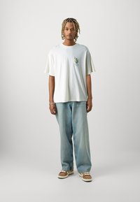 Camiseta de algodón blanca con mangas cortas y anchas, que presenta un gráfico colorido en el pecho; combinada con unos vaqueros de pierna ancha de color azul claro y zapatos marrones.