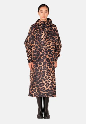 Vrouw staat frontaal, draagt een lange, losse regenjas met luipaardprint en capuchon en zwarte laarzen, tegen een effen lichte achtergrond.