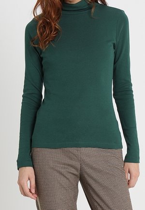 Kobieta ubrana w ciemnozielony golf z długim rękawem oraz brązowo-białe spodnie w kratę, ręce luźno opuszczone wzdłuż boków.