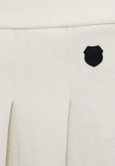Cream-colored fabric with soft texture, featuring a black embroidered shield logo and subtle pleats for design detail.