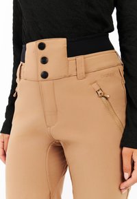 Beige skibroek met een brede zwarte elastische tailleband, drie zwarte knopen en een ritszak aan de zijkant. Soepele textuur en getailleerd ontwerp.