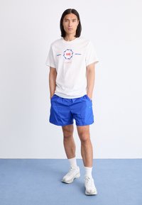 Hvid Nike t-shirt med blå og rød grafisk tekst; parret med lyseblå shorts og hvide/grå sneakers med teksturerede såler.