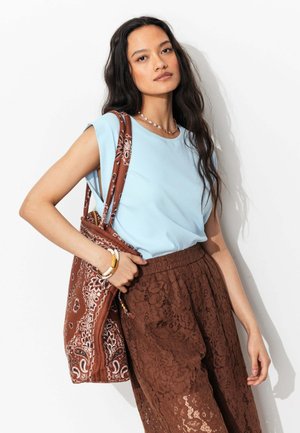 Jeune femme aux longs cheveux foncés portant un haut sans manches bleu clair, un pantalon en dentelle marron et un sac fourre-tout marron à motifs.