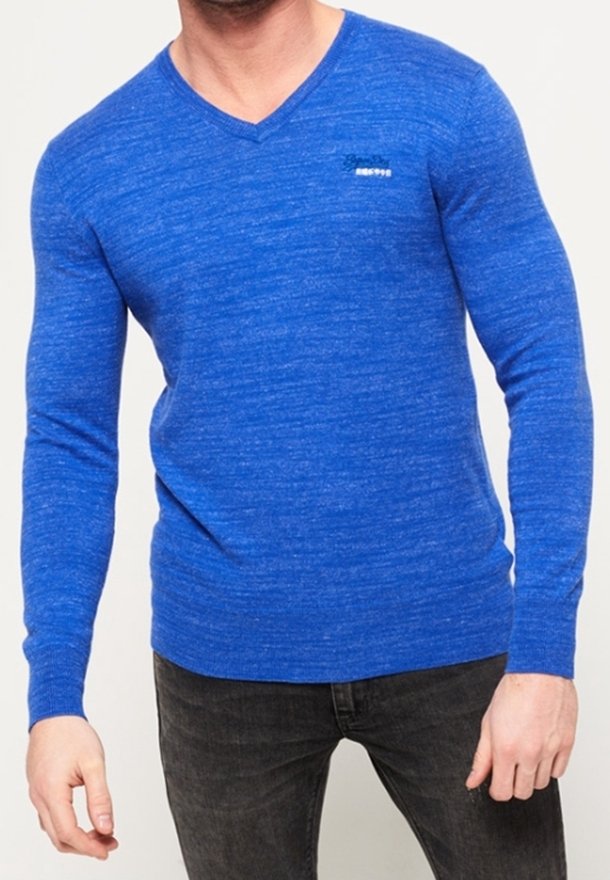 Blauer Strickpullover mit V-Ausschnitt, langen Ärmeln und strukturiertem Finish. Besitzt ein kleines Logo auf der linken Brust. Getragen mit dunklen Jeans.
