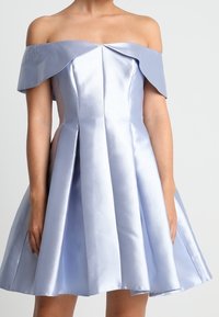 Žena v světle modrých saténových šatech s odhalenými rameny, s upnutým korzetem a rozšířenou sukní, na jednobarevném pozadí.