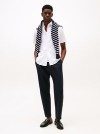 Uomo con camicia bianca a maniche corte, pantaloni blu navy, mocassini neri e un maglione a righe blu navy e bianche appoggiato sulle spalle, in piedi con le mani nelle tasche.