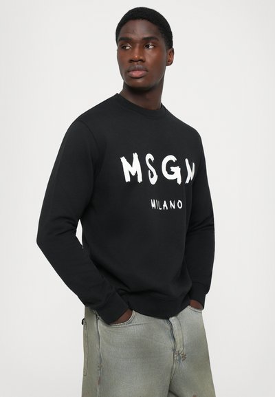 Mann in schwarzem MSGM Milano Sweatshirt und hellen Jeans, Hände in den Taschen, schaut zur Seite vor einem weißen Hintergrund.
