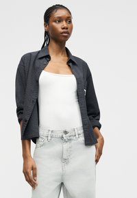 Fiatal nő fonott hajjal, sötét mintás nyitott ingben, fehér trikó és világosszürke farmernadrág felett, egyszerű háttér előtt.