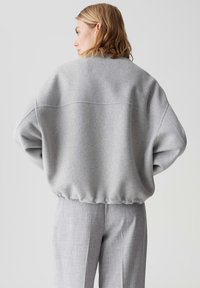 Frau mit kurzem blonden Haar, die einen oversized hellgrauen Pullover und passende graue Hosen trägt und mit dem Rücken zu einer schlichten Hintergrund steht.
